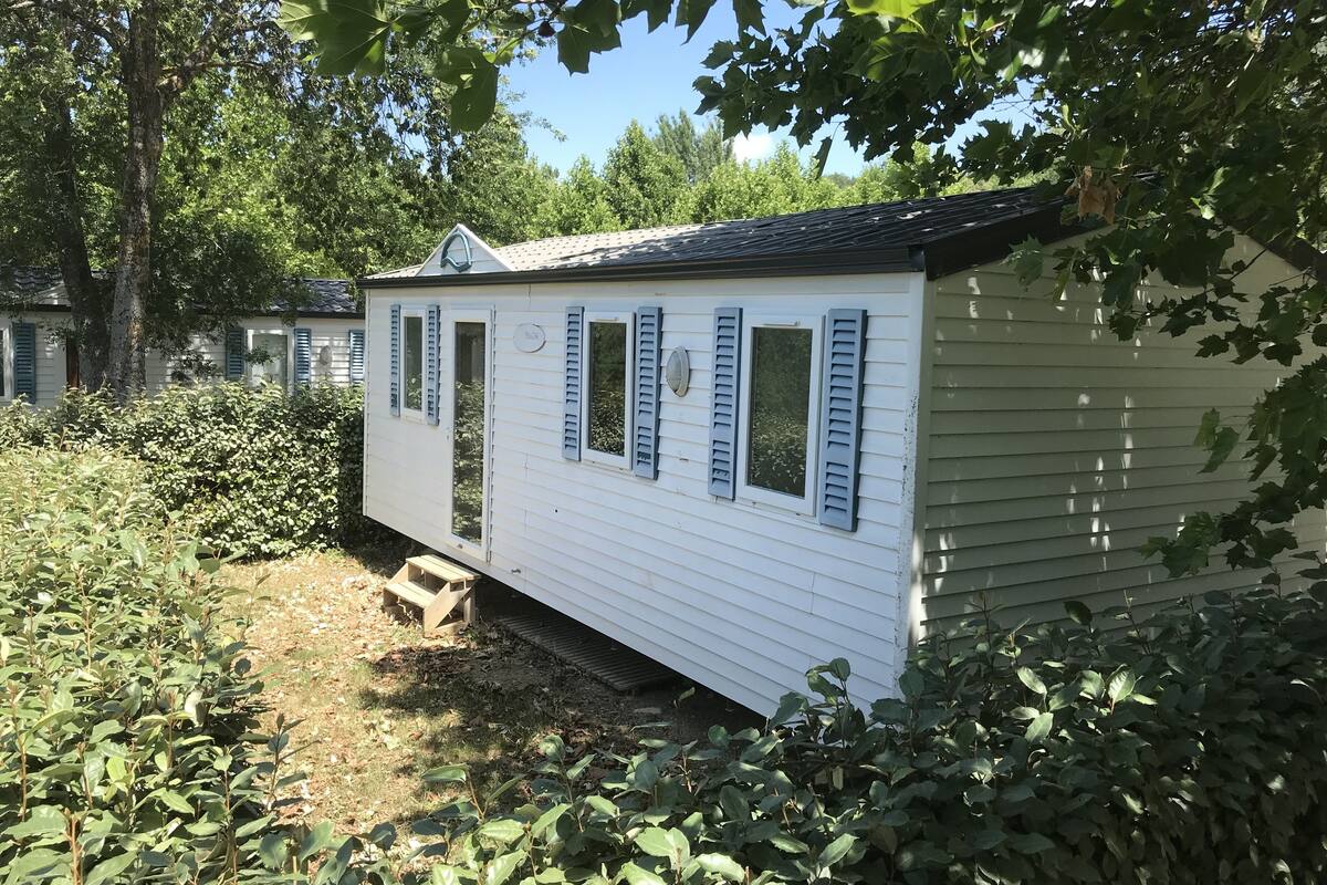 A white mobile home with blue shutters, surrounded by greenery and trees.
