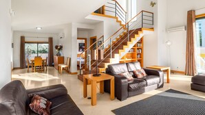 A modern living room with a staircase, leather sofas, a dining area, and a bookshelf.