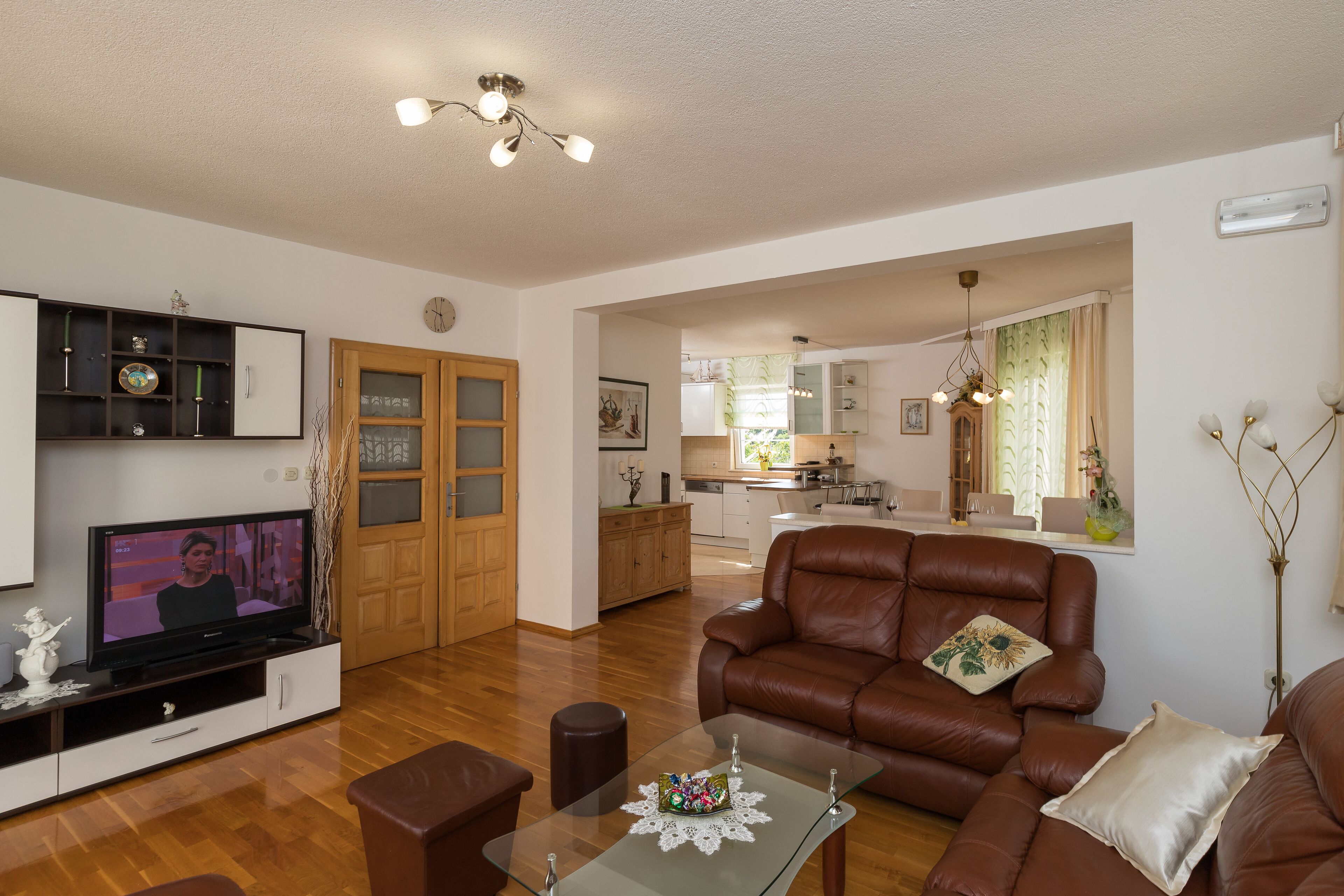 Un vaste salon comprenant un canapé en cuir, une table basse en verre et un téléviseur posé sur un meuble de rangement en bois. On aperçoit une cuisine et un coin repas à l’arrière-plan.