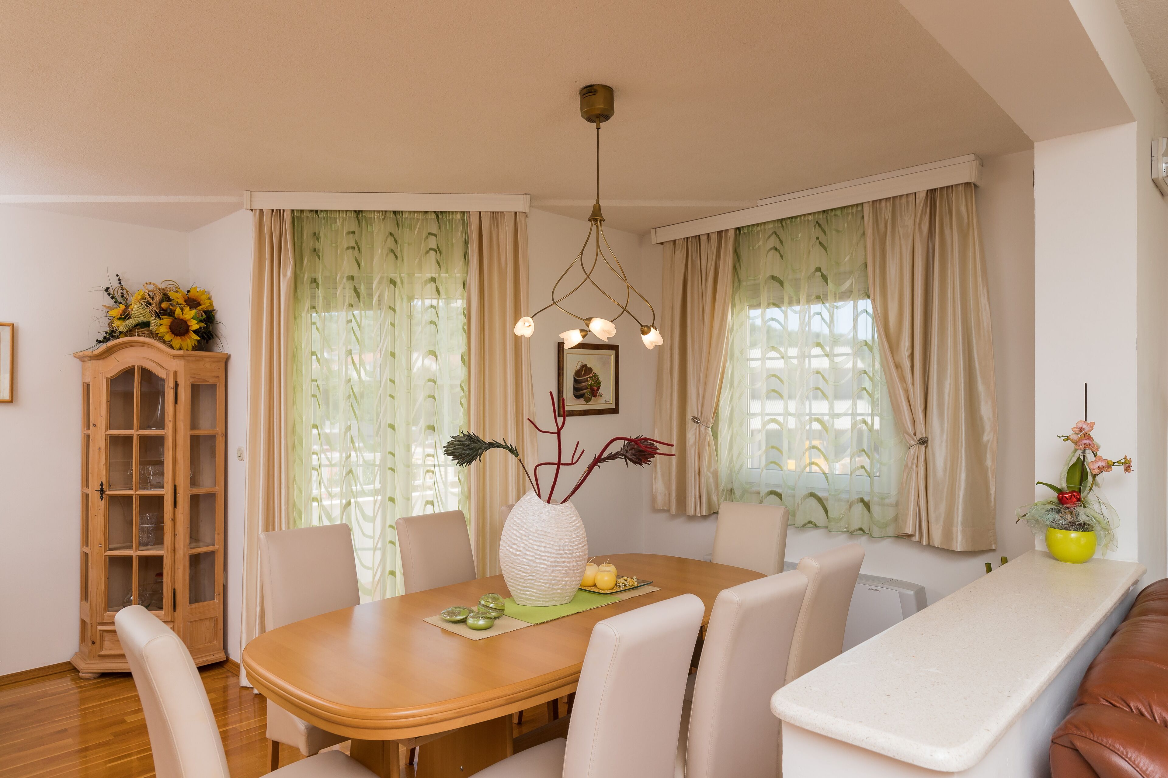 A dining area with a wooden table, white chairs, a vase with flowers, and a cabinet with glass doors.