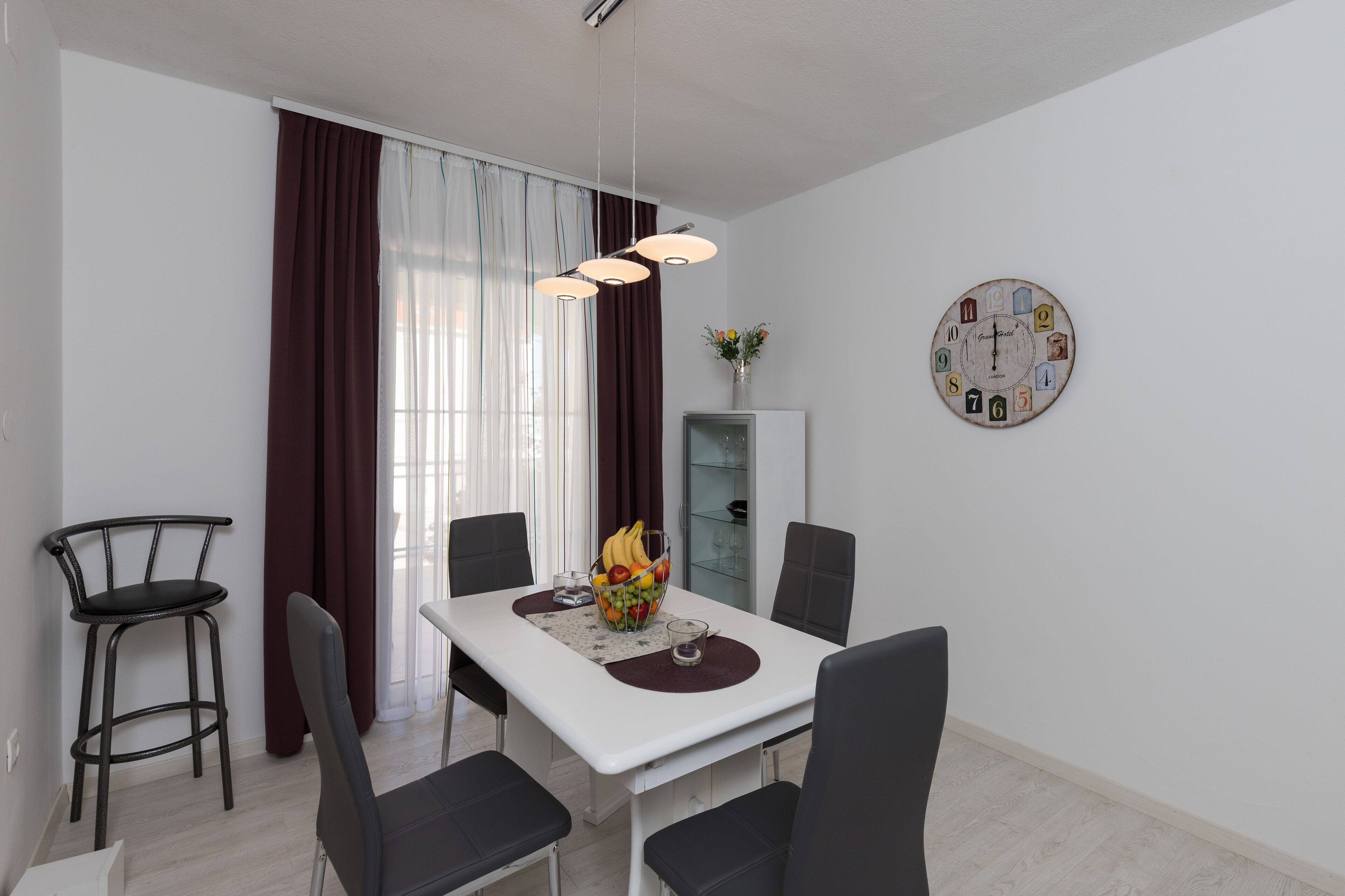 Een eethoek met een witte tafel, zwarte stoelen, een fruitschaal en een klok aan de muur.