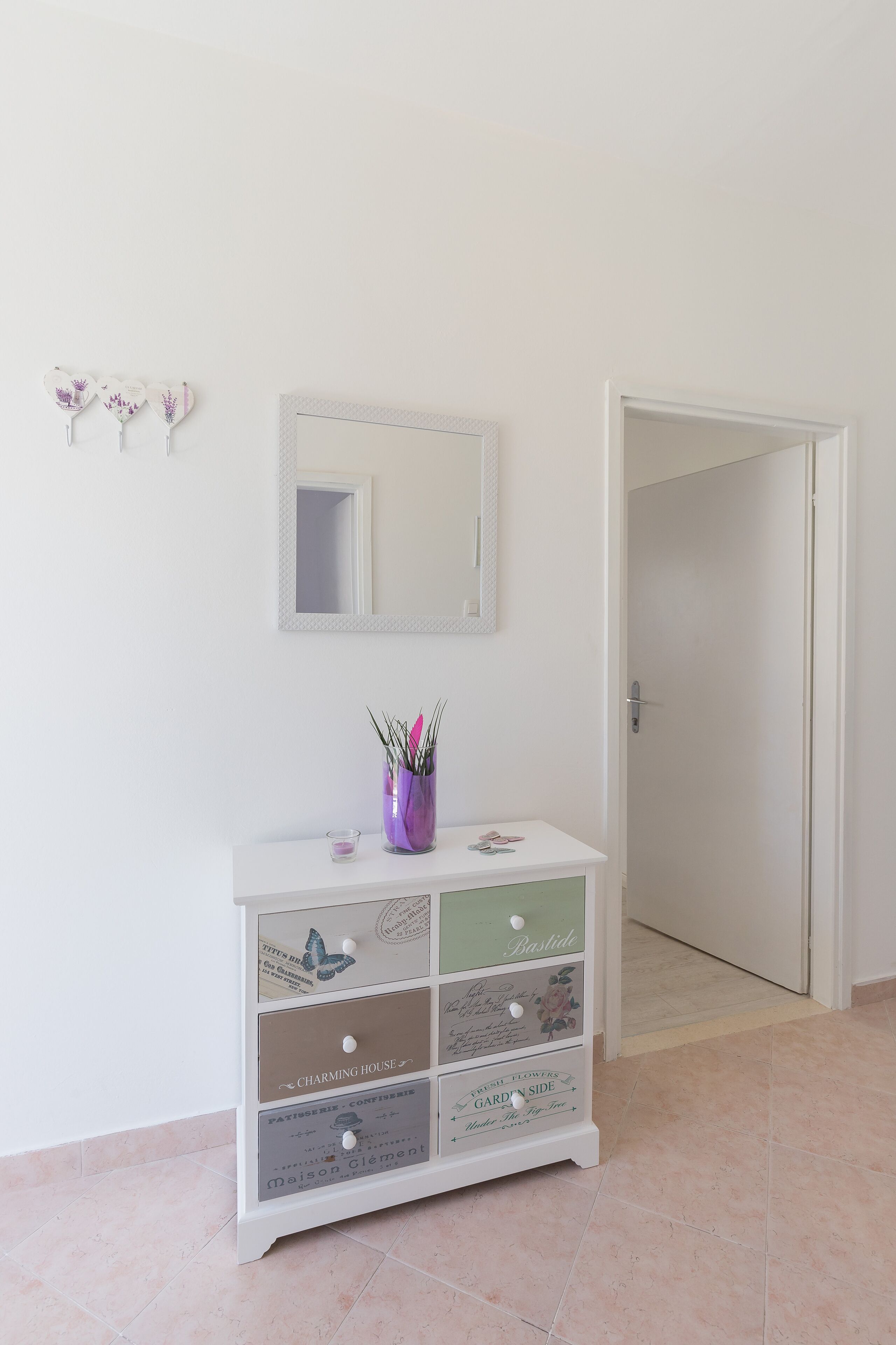 A white dresser with drawers labeled with names like "Charming House" and "Garden Side." A mirror hangs above the dresser, and a door is visible to the right.
