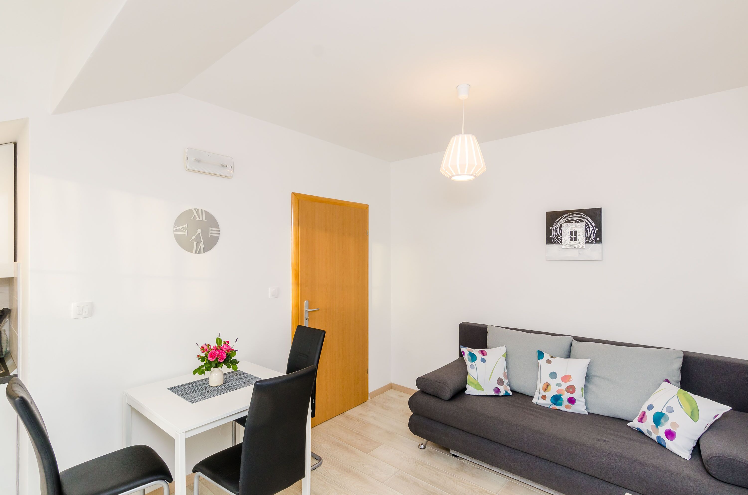 A modern living room with a grey sofa, a dining table with two chairs, a wooden door, and a wall clock.