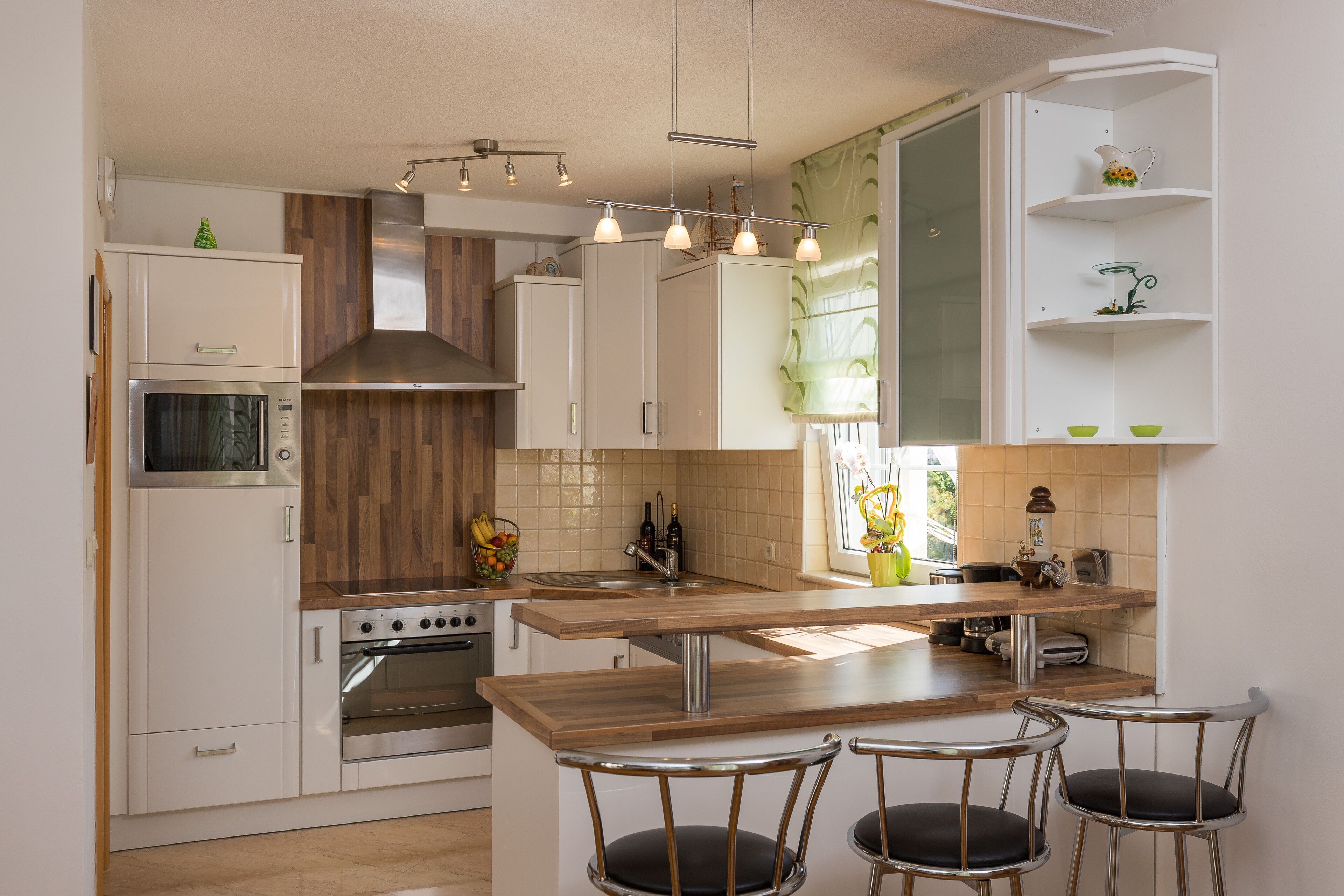 Una cocina moderna con armarios blancos, encimera de madera y horno empotrado. Hay tres taburetes de bar y una ventana con vistas a zonas verdes.