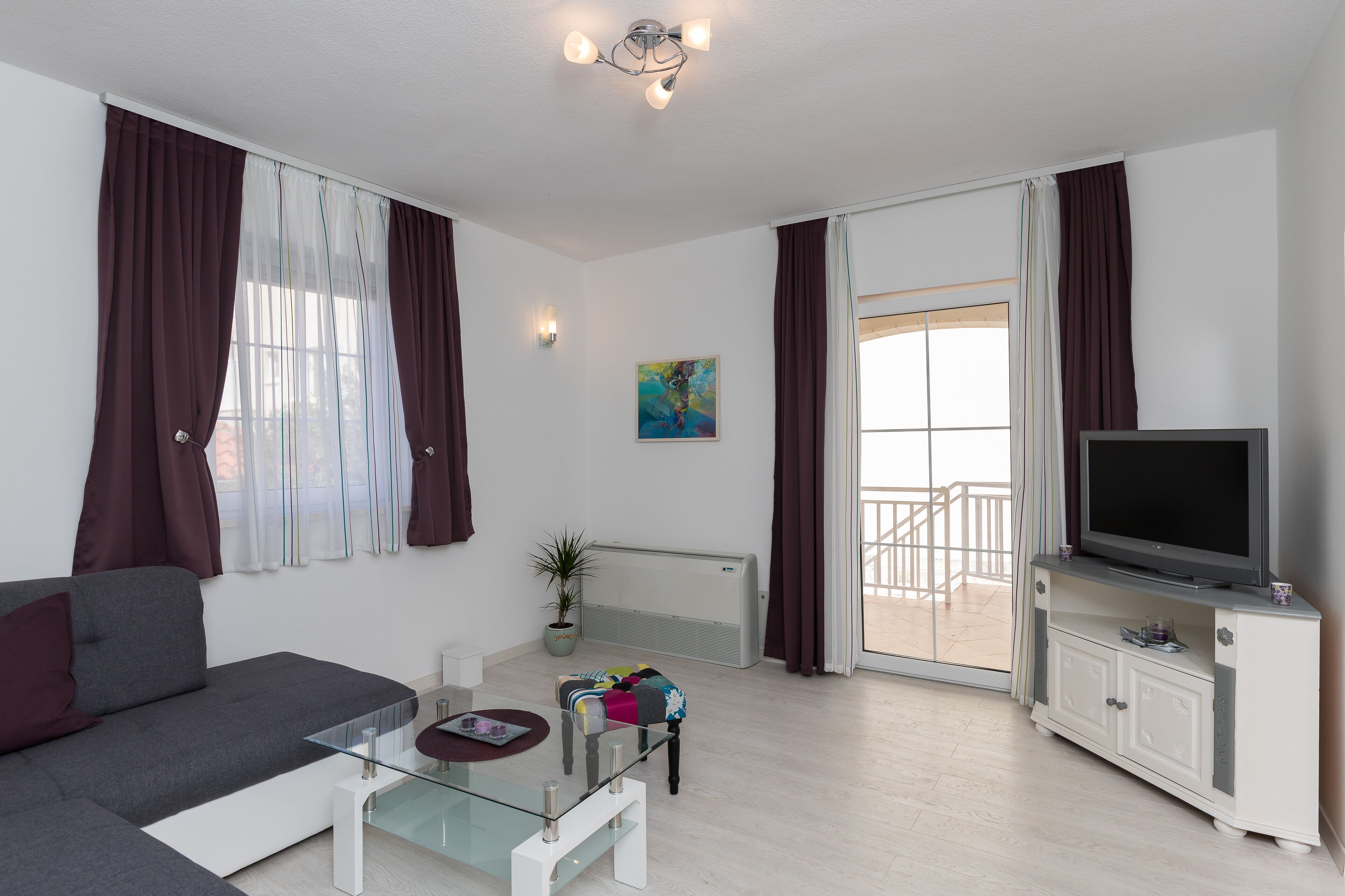 A modern living room with a sofa, a glass coffee table, a flat-screen TV on a cabinet, and a balcony door.