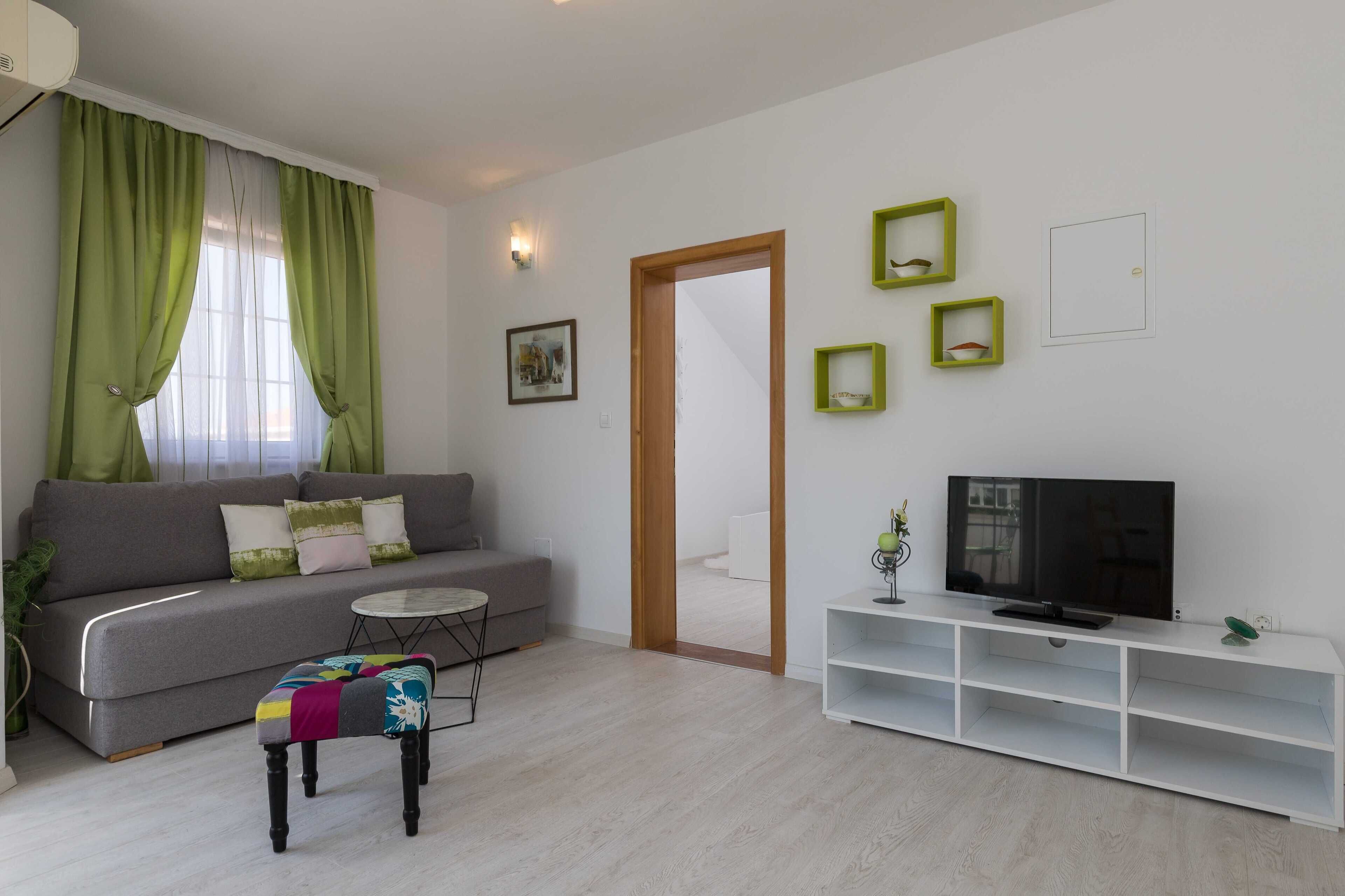 Un soggiorno moderno con un divano grigio, un tavolino rotondo, un poggiapiedi colorato, una TV su un mobile bianco e quadri con cornice verde alle pareti.