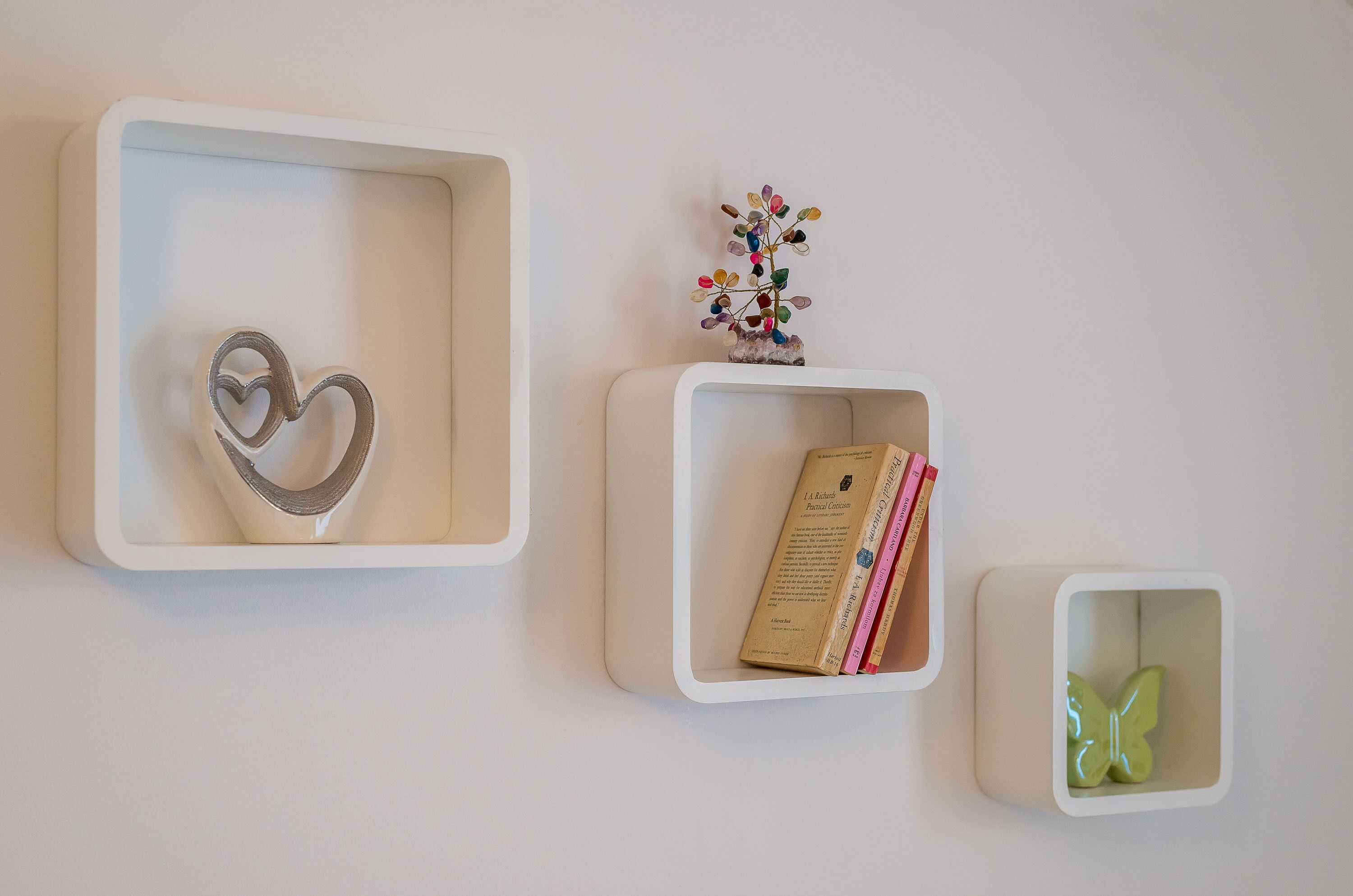 Tres repisas blancas montadas en la pared con un objeto con forma de corazón, un pequeño árbol decorativo y libros.