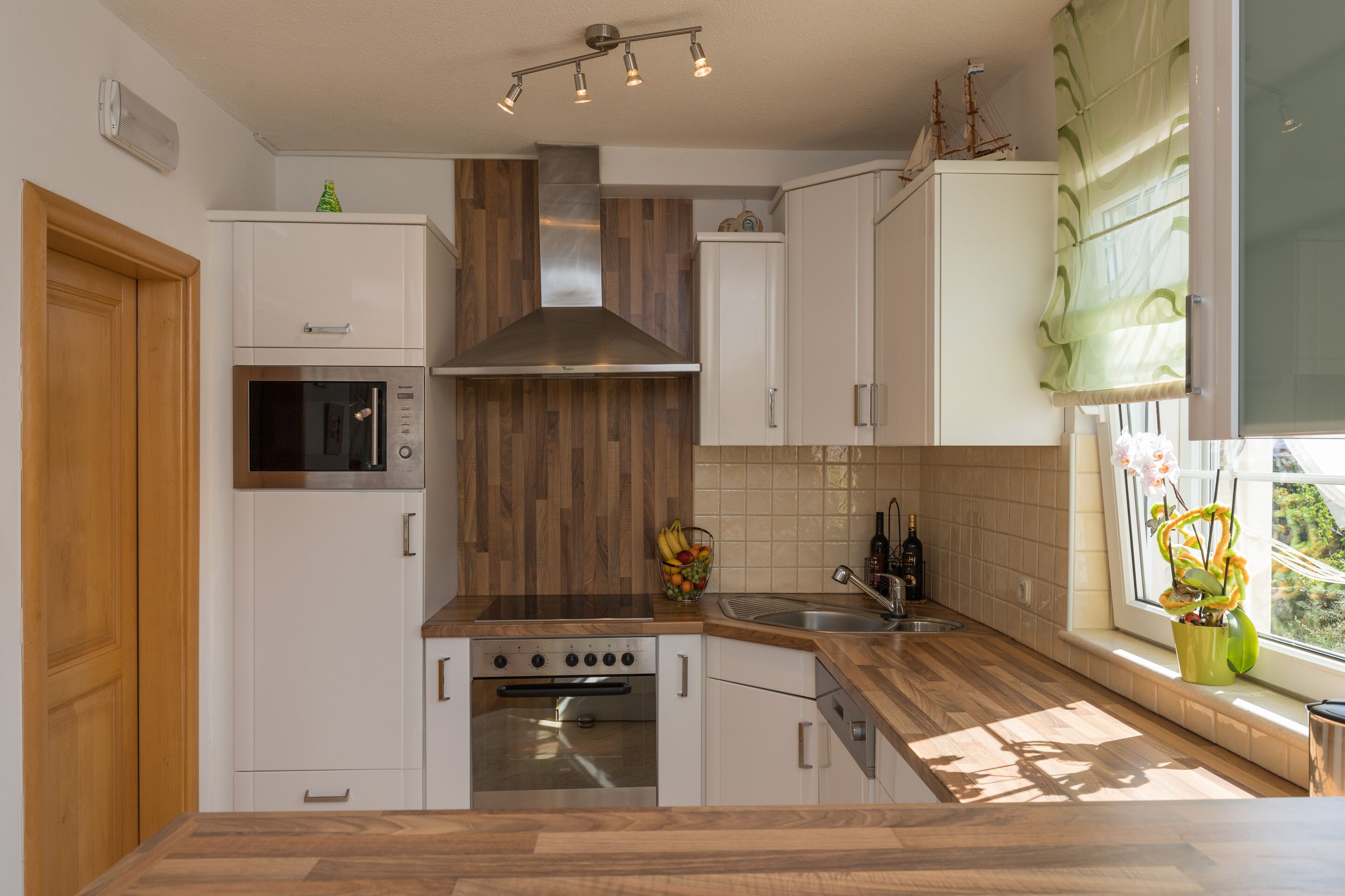 A modern kitchen with white cabinets, a stainless steel range hood, a built-in oven, a microwave, and a window with a view of greenery.