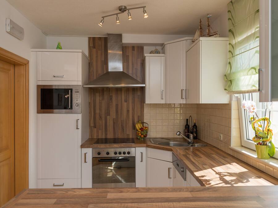 A modern kitchen with white cabinets, a stainless steel range hood, a built-in oven, a microwave, and a window with a view of greenery.