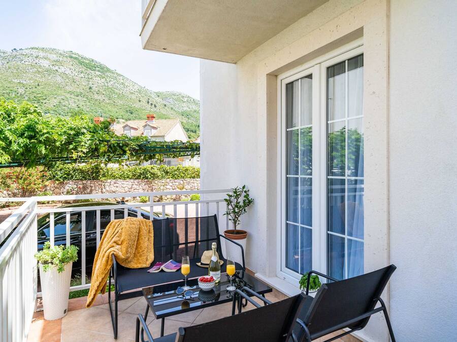 A balcony with a table set for two, a potted plant, and a view of a mountain and houses in the background.