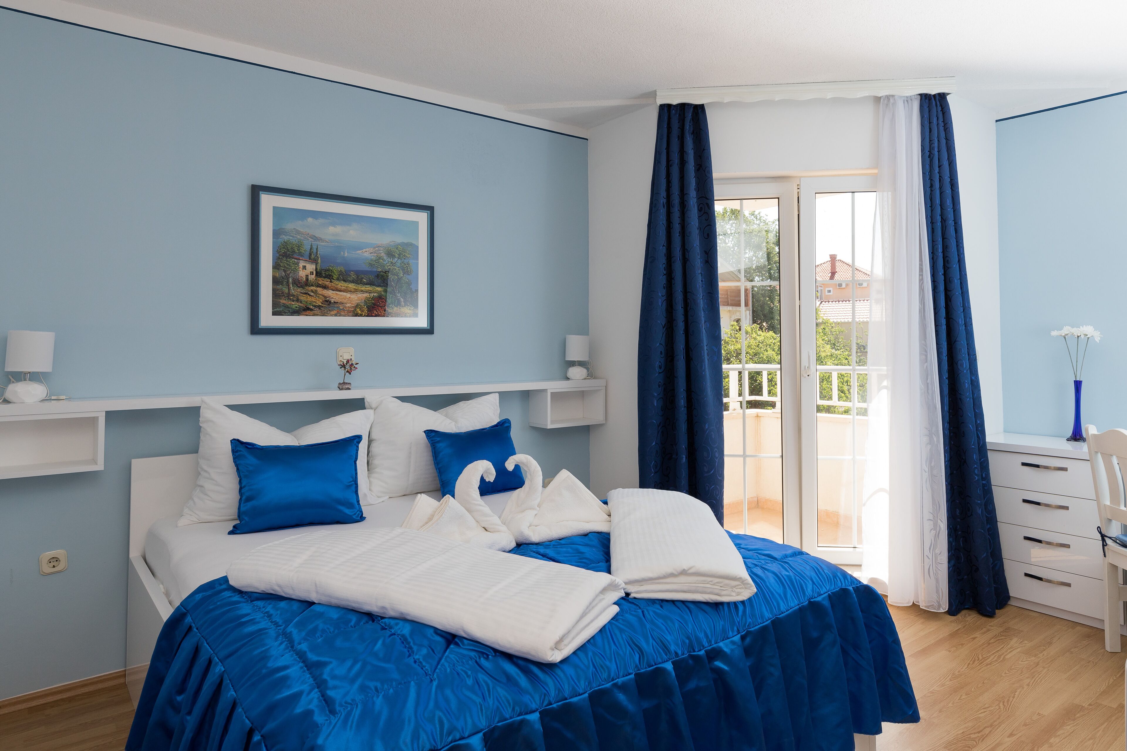 Ein Schlafzimmer mit Bett, blauer Bettwäsche, einem gerahmten Bild an der Wand, einem Fenster mit Vorhängen und einem Balkon mit Aussicht.