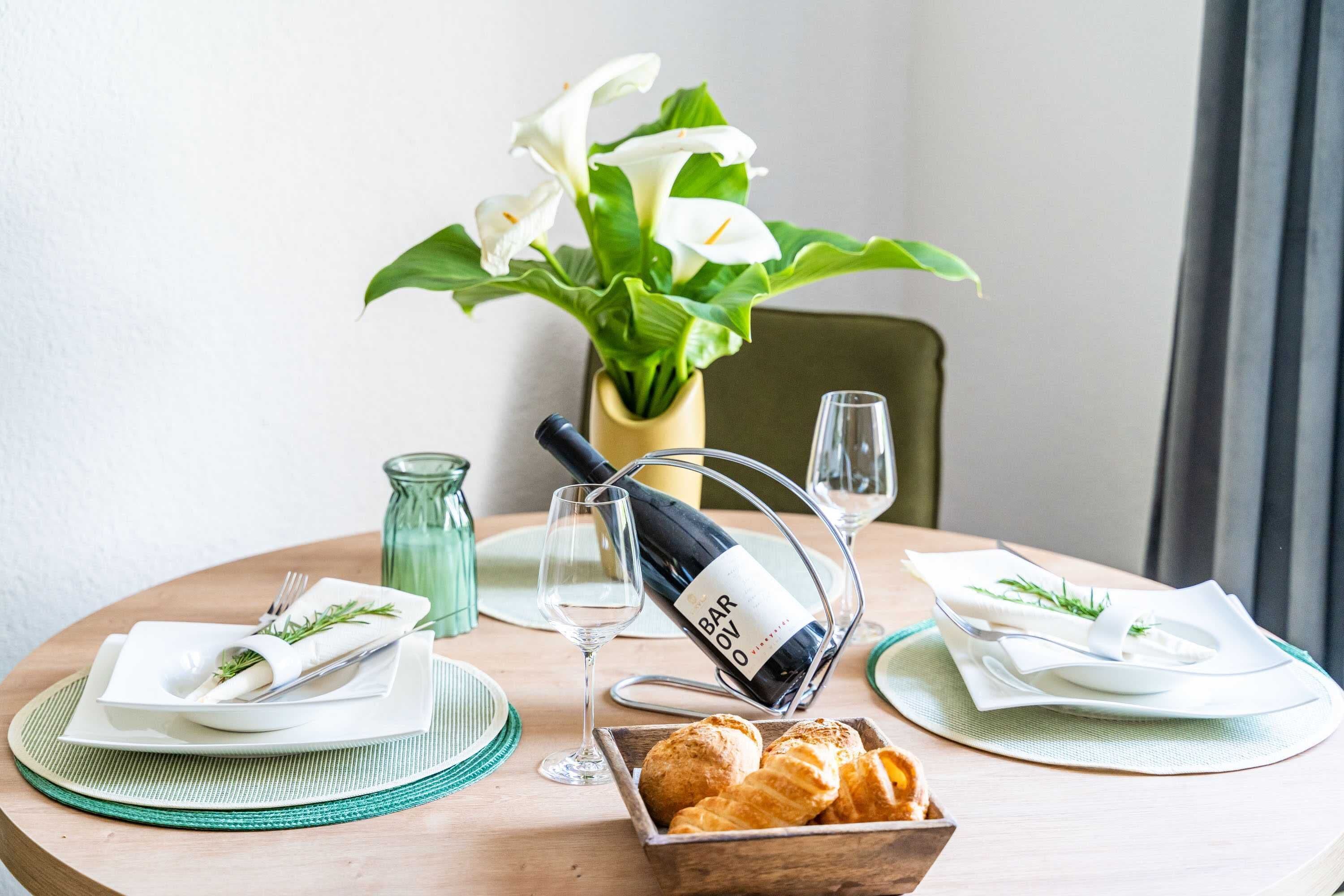 Una mesa de comedor con una botella de vino Barolo, dos copas, un bol de cruasanes y un jarrón con flores blancas.