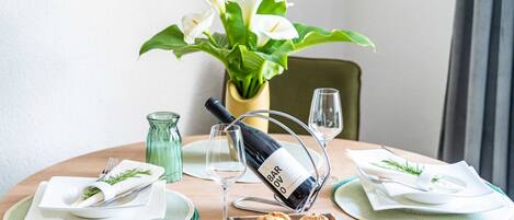 Una mesa de comedor con una botella de vino Barolo, dos copas, un bol de cruasanes y un jarrón con flores blancas.