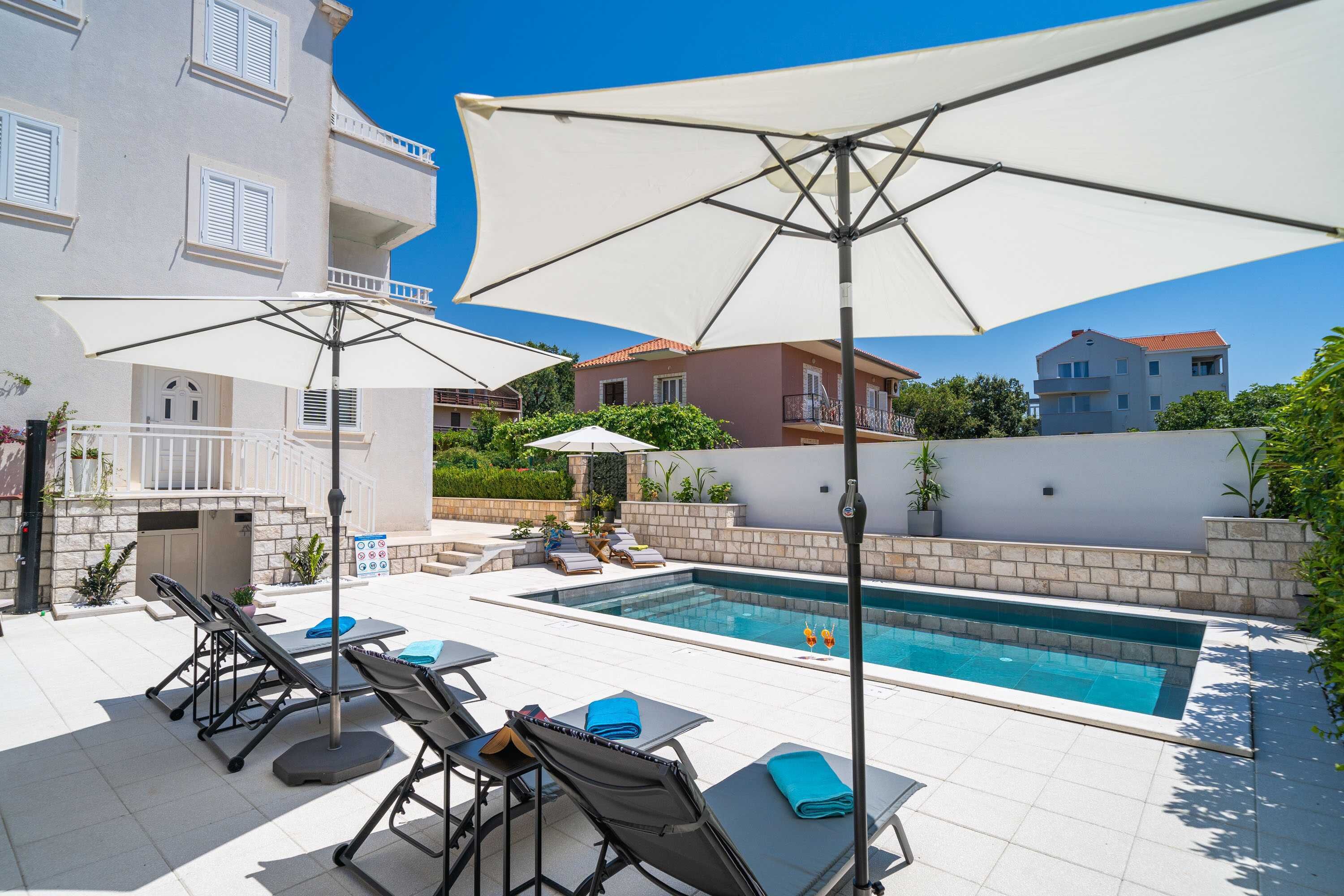 A pool area with sun loungers and umbrellas, a pool, and a building with balconies.