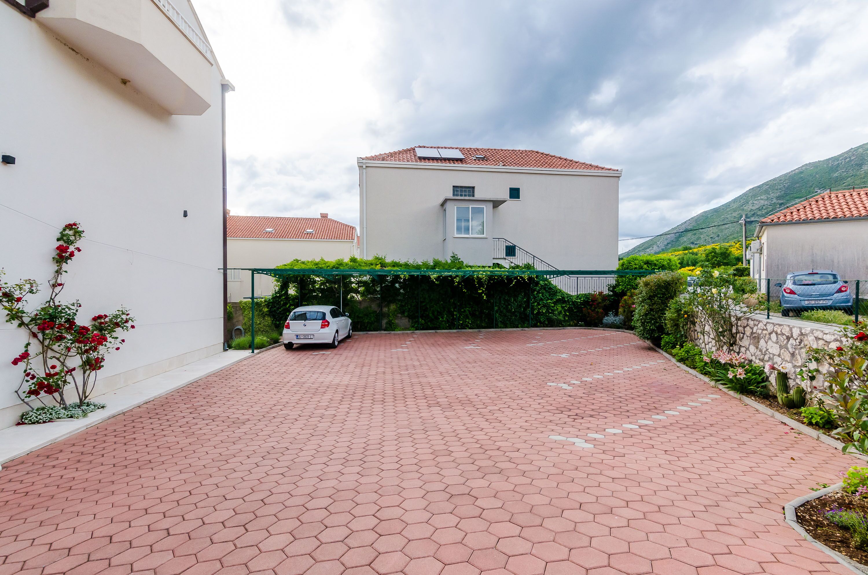 A paved driveway leading to a building with a red-tiled roof, surrounded by greenery and a parked car.