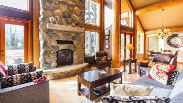 A cozy living room with a stone fireplace, wooden beams, large windows, and comfortable seating.
