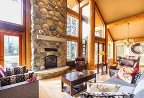 A cozy living room with a stone fireplace, wooden beams, large windows, and comfortable seating. - Large Family Chalet on Nicklaus North Golf Course (Whistler)