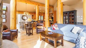 A spacious living area with a sofa, coffee table, and leather armchair. A dining area with a chandelier and wooden beams is visible in the background.