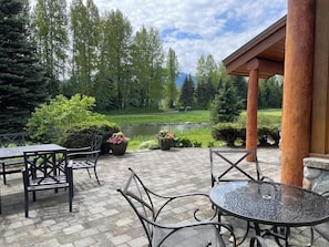 An outdoor patio with tables and chairs, a pond, and a forested backdrop. - Large Family Chalet on Nicklaus North Golf Course (Whistler)