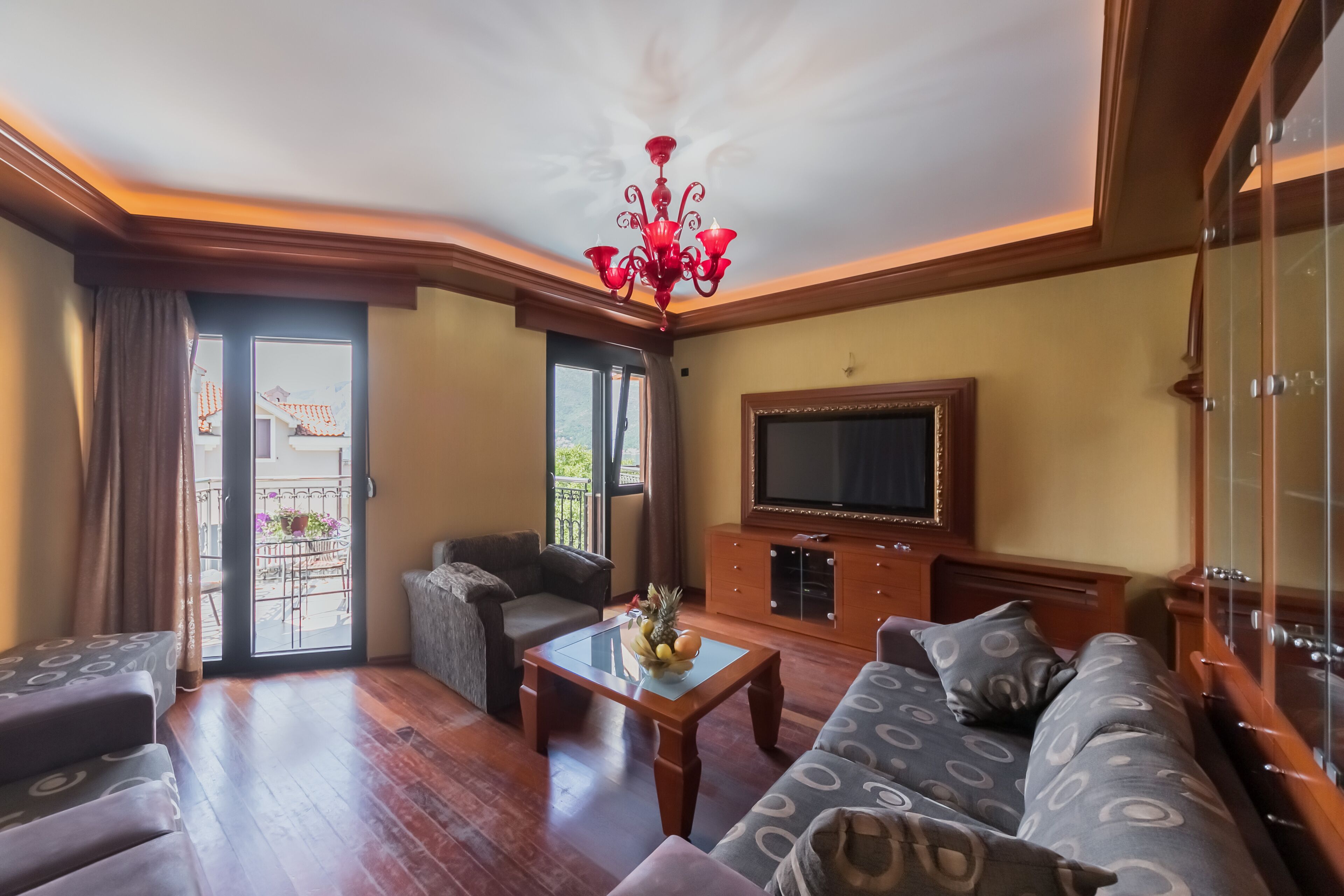 A living room with a wooden coffee table, a sofa, a television on a wooden cabinet, and a balcony with a view of the mountains.