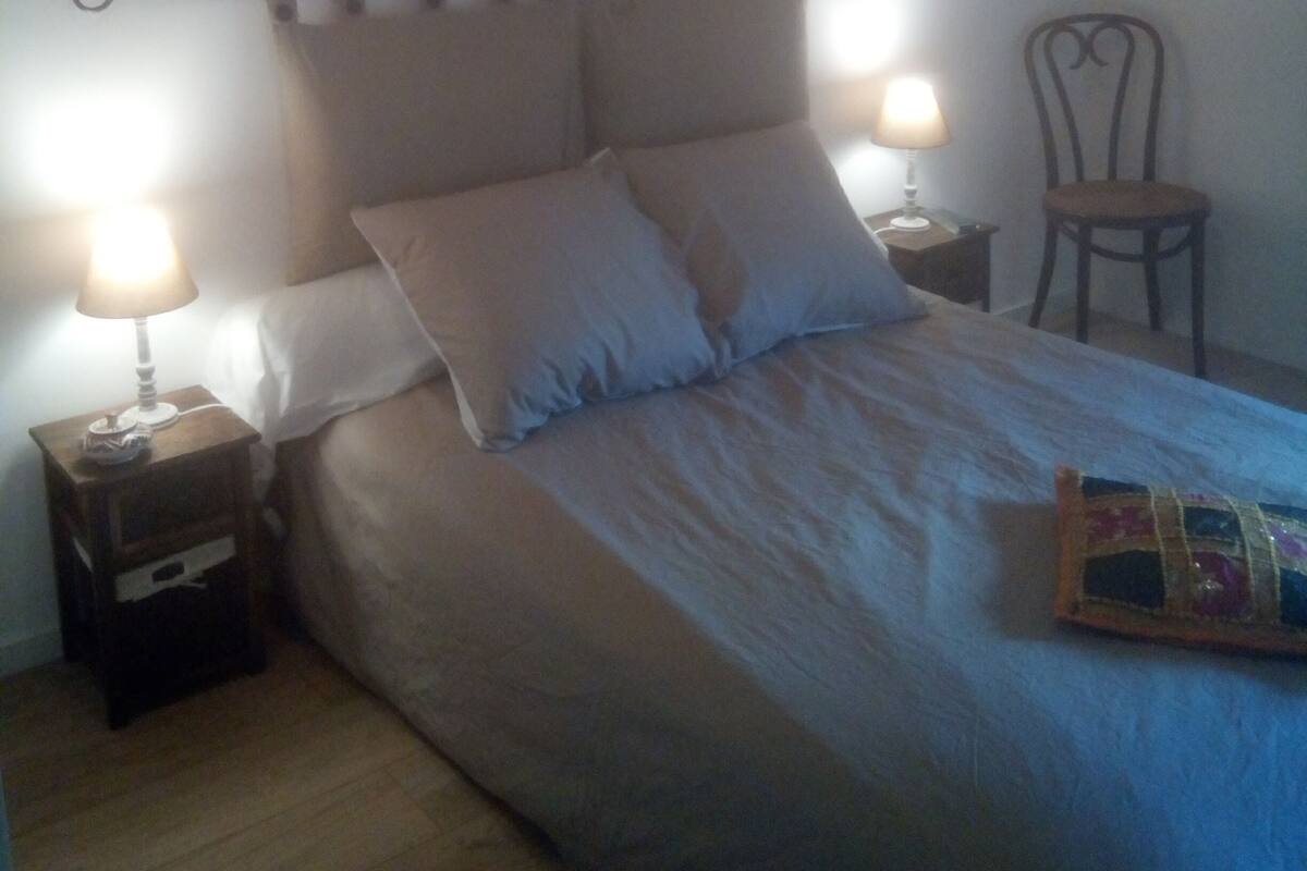 Une chambre à coucher comprenant un lit, deux lampes de chevet, une chaise, une œuvre d’art accrochée au mur et un plafond en bois.