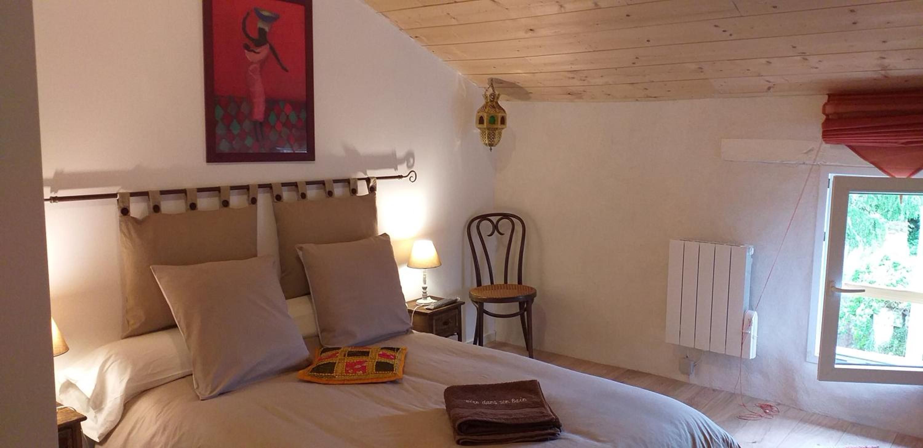 Une chambre à coucher avec un lit, des oreillers, une table de chevet, une chaise, une lampe fixée au mur et une fenêtre donnant sur un beau paysage.