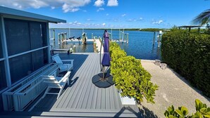 Een houten terras met lounge fauteuils en een parasol, met uitzicht op een rustig waterlichaam met steigers in de verte.