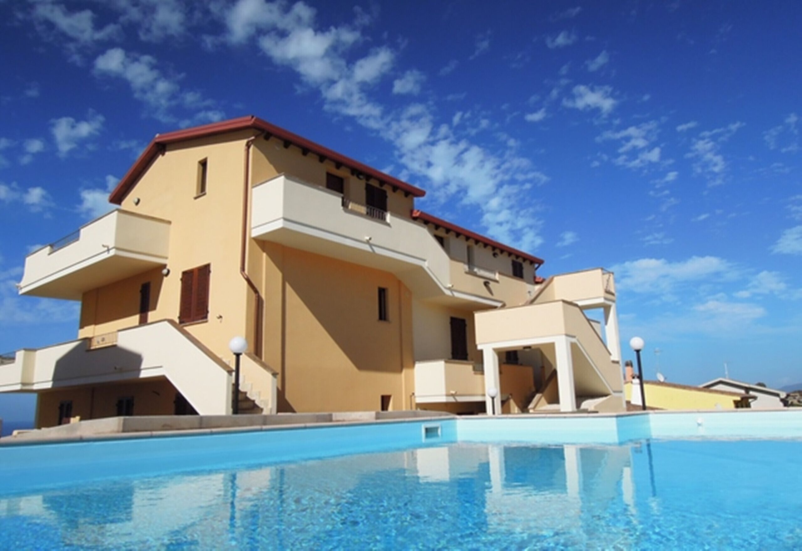 A multi-story hotel with a swimming pool in front, featuring balconies and a red roof.