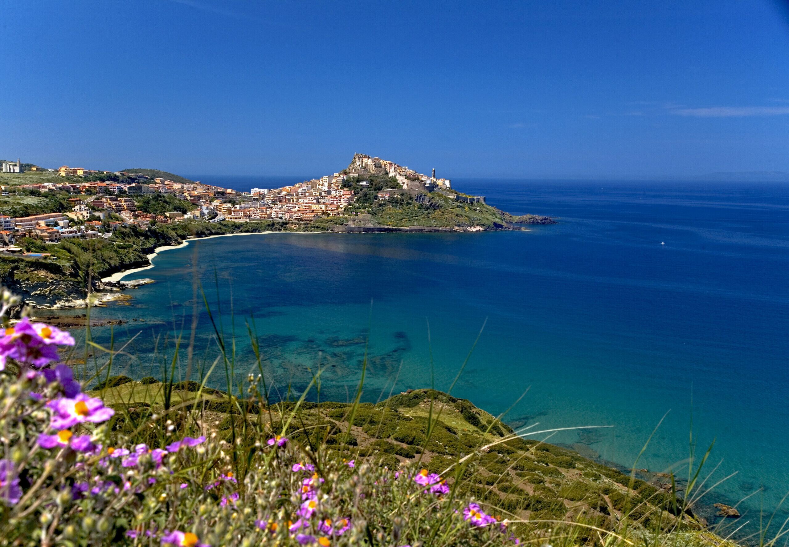 A coastal town with a prominent hilltop structure, clear blue sea, and surrounding greenery.