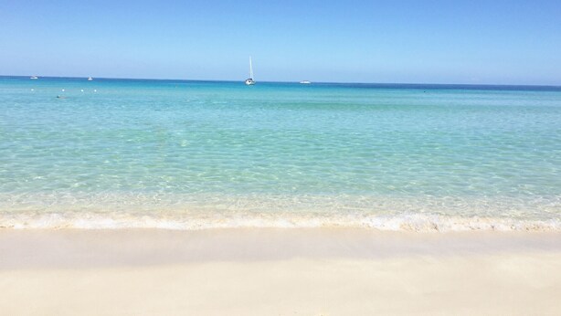 Ein Strand mit klarem türkisblauem Wasser, einem sandigen Ufer und einem Segelboot in der Ferne.
