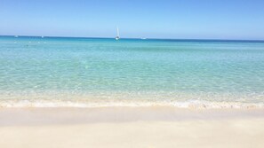 Une plage à l’eau turquoise et limpide, un rivage sablonneux et un voilier à l’horizon.