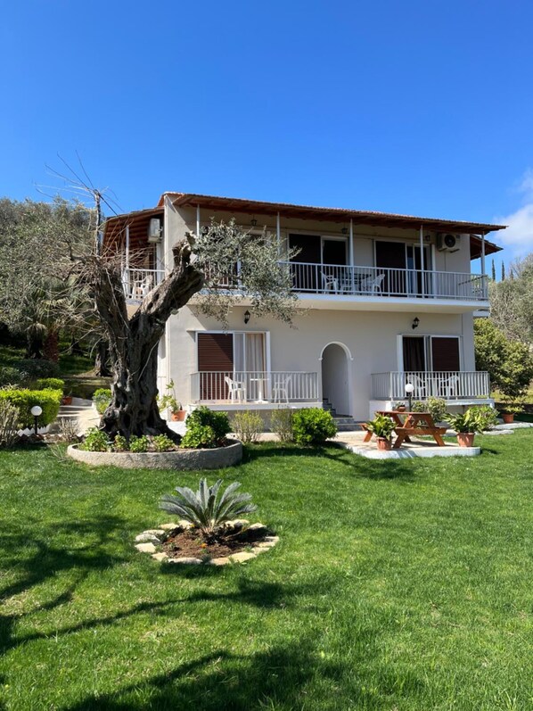 Una casa de dos pisos con balcón, rodeada por un jardín cuidado con diversas plantas y un árbol grande.