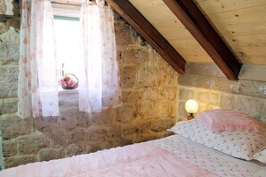 Een slaapkamer met een stenen muur, een houten plafond, een bed met roze beddengoed en een raam met bloemen gordijnen.