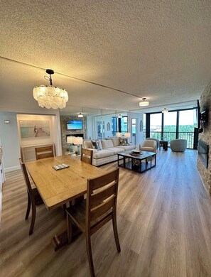 A spacious living room with a wooden dining table, sofas, and a large window offering a view. - Siesta Key Beach Condo w/ water view & pool, half mile to the beach! (Siesta key)
