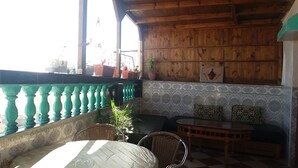 A cozy patio area with a wooden ceiling, a green balustrade, a tiled wall with a decorative pattern, a wicker chair, a cushioned bench, and potted plants. - Bassi Apartment (Taghazout)