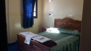 A bedroom with a wooden bed, a nightstand, a window with blue curtains, and a wall socket. - Bassi Apartment (Taghazout)