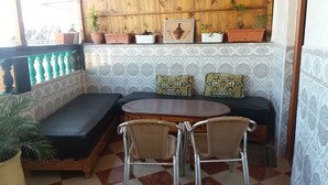 A corner seating area with a sofa and two chairs, a small table, and potted plants on a wooden shelf. - Bassi Apartment (Taghazout)