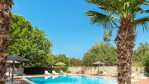 Una piscina con sillones de descanso y sombrillas, rodeada de palmeras y un muro de piedra.