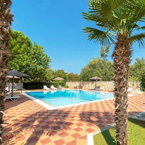 Piscina com espreguiçadeiras e guarda-sóis, cercada por palmeiras e um muro de pedra.