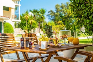 Mesa de jantar ao ar livre com diversos alimentos e bebidas, rodeada por cadeiras de madeira.