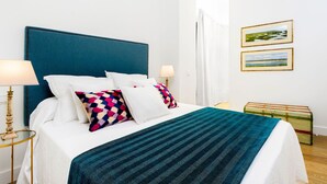 A bed with a blue headboard, white bedding, and a colorful pillow. There are two bedside lamps, a window with white curtains, and a small table with a lamp. - Genteel Home Mateos Gago II (Seville)