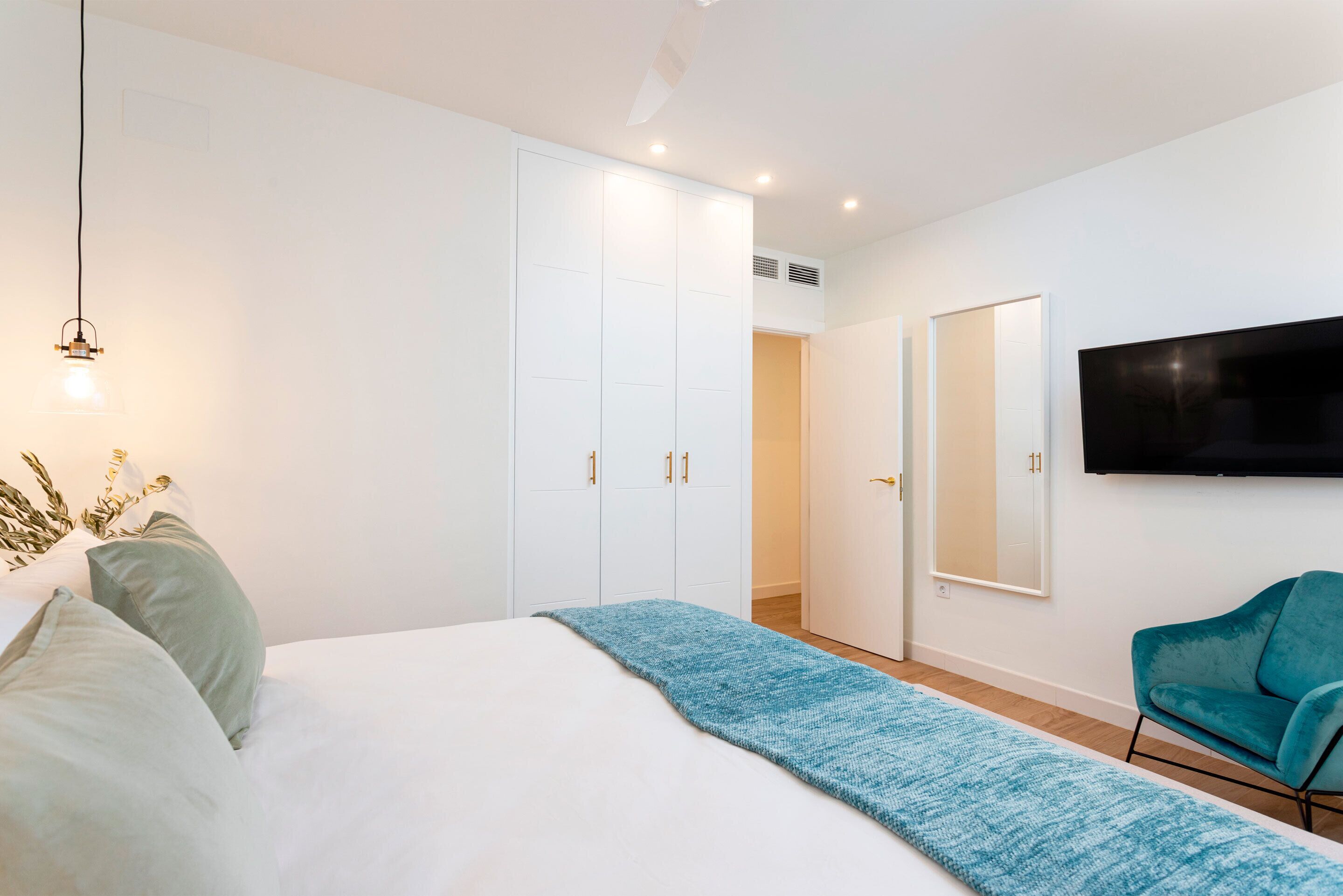 A modern bedroom with a bed, a blue throw blanket, a teal chair, and a TV mounted on the wall.
