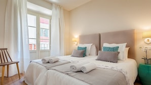 A neatly made bed with pillows and a blanket, a wooden chair, and a bedside table with a lamp and a plant.