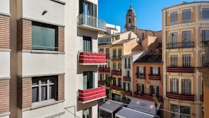 A narrow street with multi-story buildings, balconies with red curtains, and a church tower in the background.