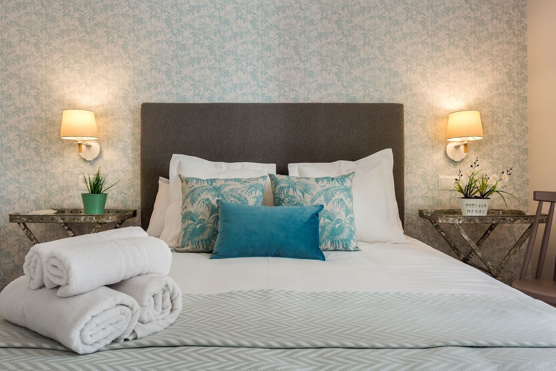 A neatly made bed with white linens and a teal pillow, flanked by two nightstands with lamps and potted plants.
