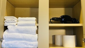 A wooden cabinet with shelves containing white towels, a black hairdryer, and two rolls of toilet paper.