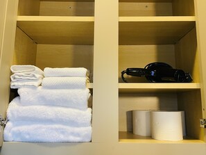 A wooden cabinet with shelves containing white towels, a black hairdryer, and two rolls of toilet paper. - Newly Renovated, Spacious Suite with Every Luxury (Richmond)