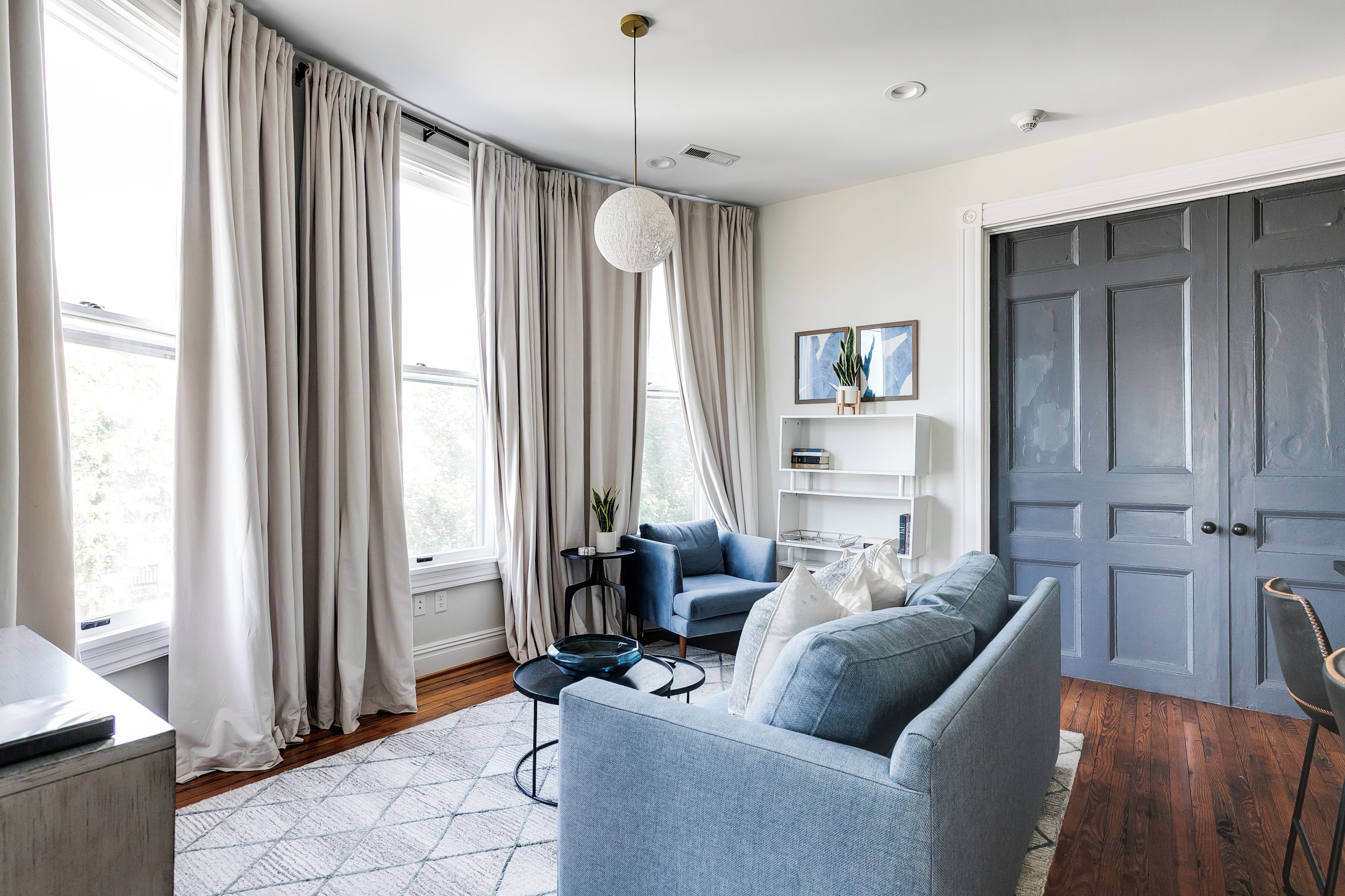 Una sala de estar moderna con un sofá, una mesita auxiliar y una silla. Hay una puerta con un panel decorativo y una ventana con cortinas.