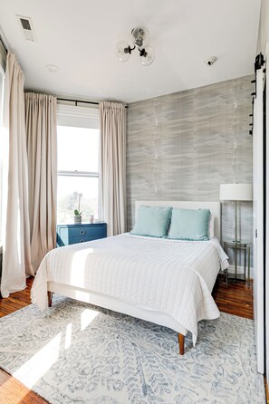 A bedroom with a bed, curtains, a nightstand, a lamp, and a window. - Newly Renovated, Spacious Suite with Every Luxury (Richmond)