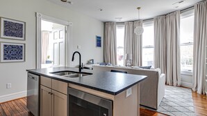Una cocina moderna con lavavajillas integrado, un lavabo y una isla grande. Hay una ventana con cortinas, un sofá y una alfombra en el suelo.